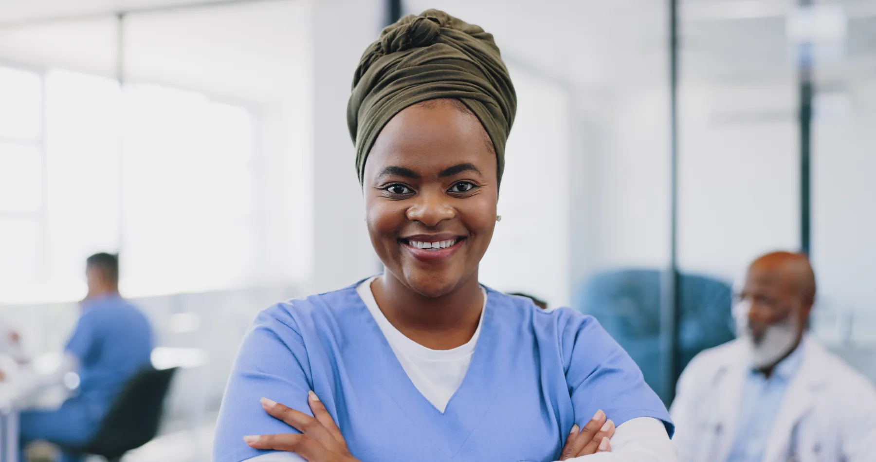 A smiling woman in pale blue scrubs standing indoors with her arms crossed.