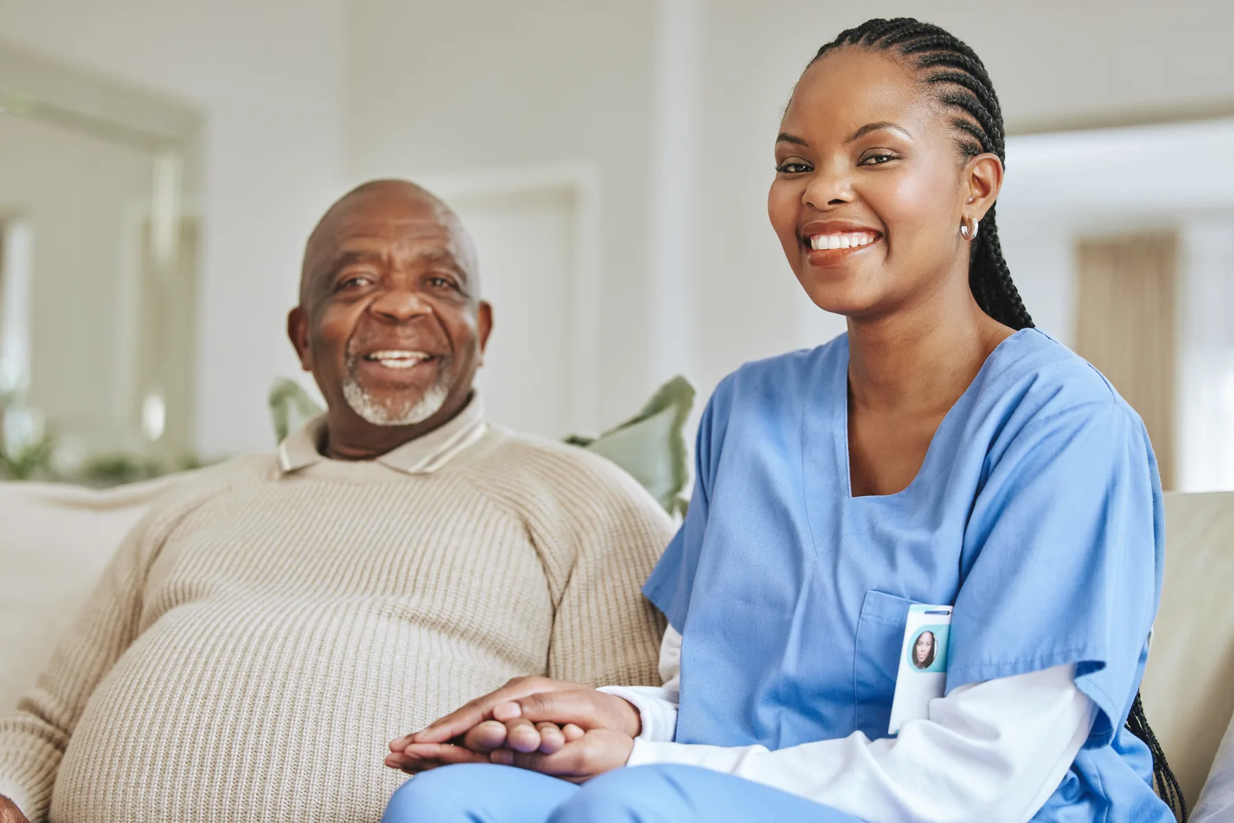 An older man seated at home beside a smiling woman in blue scrubs.